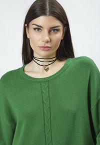Grüner Strickpullover mit rundem Halsausschnitt und vertikalem Zopfstrickdetail. Getragen mit einem schwarzen Choker mit einem Herzanhänger.