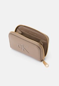 Beżowy skórzany portfel ze złotym suwakiem. Posiada wytłoczone logo "CK", wiele miejsc na karty oraz gładką teksturę. Kompaktowy prostokątny kształt.