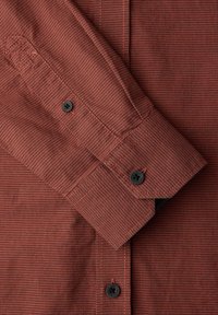 Chemise à manches longues rouge-brun avec un fin motif à carreaux noirs, dotée de boutons noirs sur le poignet et la patte de manche.