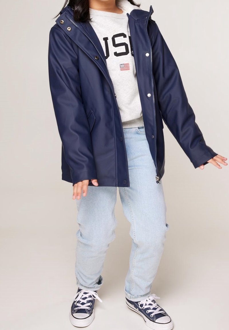 Enfant portant un imperméable bleu marine, un jean clair, des baskets bleu marine et un sweat-shirt gris avec "USA" et un petit drapeau.