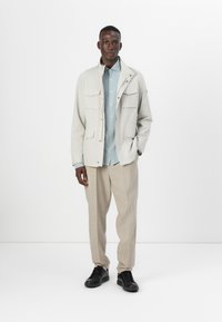 Light gray lightweight jacket with multiple pockets, worn over a pale blue shirt and beige trousers. Black sneakers complete the outfit.