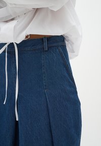 Denim hlače z visokim pasom, naborki in modro barvo. Oblikovanje vključuje zavezovanje pasu in kontrastne šivantske podrobnosti.