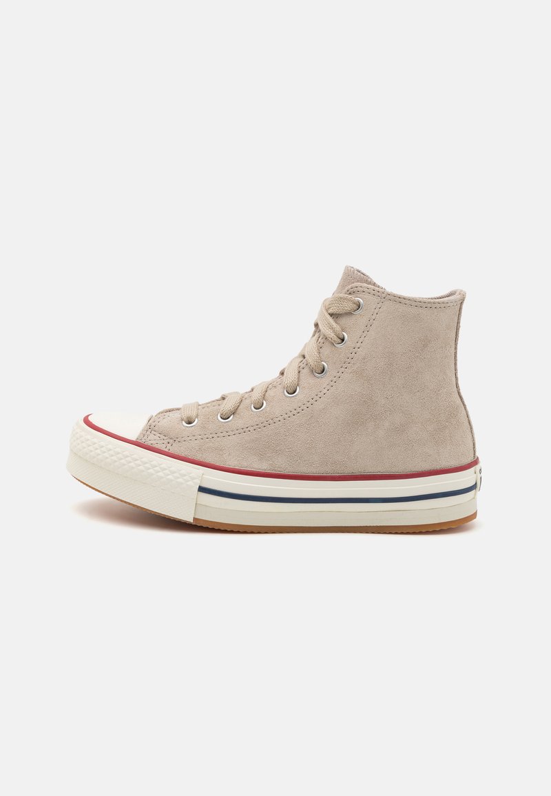 Beige suede high-top sneaker with white rubber toe cap, red and blue striped midsole, and six eyelets for laces. Textured surface.