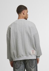 Pulover gri tricotat cu torsade, cu manșete și tiv în dublu. Presentând o etichetă decorativă în partea stângă jos. Asortat cu blugi rupți.