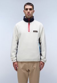 Fleece pullover i creme med marineblå detaljer og høj krave. Har en rød lynlås og et mærketikette på brystet.