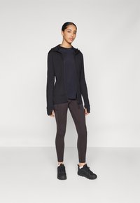 Melns zip-up džemperis ar kapuci, saskaņots ar tumši brūnām legingām. Iezīmē garas piedurknes ar īkšķu caurumiem un melnām sporta kurpēm.