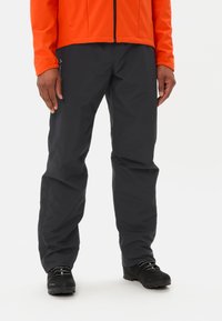 Pantalons étanches noirs en tissu léger avec une texture lisse. Portés avec une veste orange vif et des bottes noires.