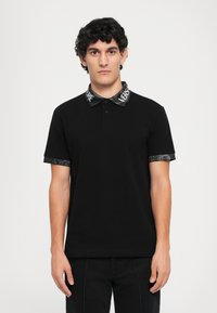 Sort polo shirt med en knaplukning, der har en mønstret krave og manchetter med ordet "VERSACE" i hvidt. Glat stof og slim fit.