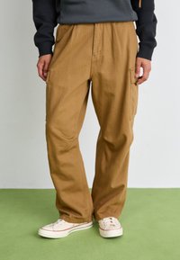 Beige cotton cargo pants with a loose fit and elastic waistband, featuring side pockets. Paired with white sneakers with red accents.