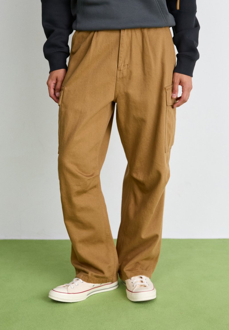 Beige cotton cargo pants with a loose fit and elastic waistband, featuring side pockets. Paired with white sneakers with red accents.