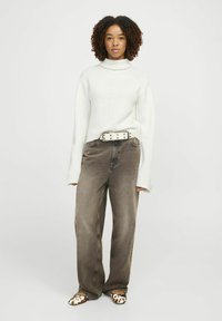 Pull à col roulé blanc côtelé, pantalon en denim brun large, ceinture blanche avec un matériel circulaire, et chaussures à enfiler à motifs avec des accents de motifs animaliers.