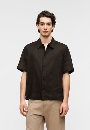 Kurzärmeliges Hemd aus dunkelbraunem Leinen mit spitzem Kragen, geradem Saum und dezentem Strukturmuster, kombiniert mit hellbeigen Hosen.