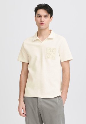 Młody mężczyzna o ciemnych włosach, ubrany w kremową koszulkę polo z krótkim rękawem, z fakturowaną kieszenią, oraz szare spodnie, stojący na białym tle.