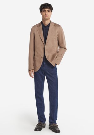 Beiger Blazer mit zwei Knöpfen, Reverskragen, kombiniert mit einem marineblauen Poloshirt und dunkelblauen Jeans, getragen mit schwarzen Schuhen, vor einem weißen Hintergrund.