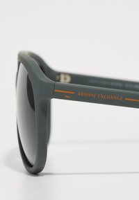 Ochelari de soare din plastic verde, cu lentile închise, având un logo portocaliu "Armani Exchange" pe braț și o finisare netedă, mată.