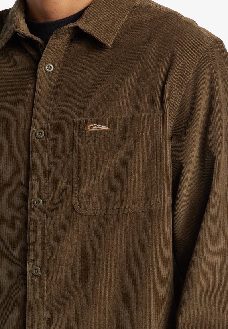 Camicia in corduroy marrone con colletto a punta, taschino frontale e piccolo logo decorativo. Il tessuto ha una finitura morbida e testurizzata.