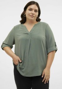 Blouse légère vert olive à col en V, avec des manches retroussées, en tissu texturé et à coupe décontractée, légèrement rentrée dans un pantalon noir.
