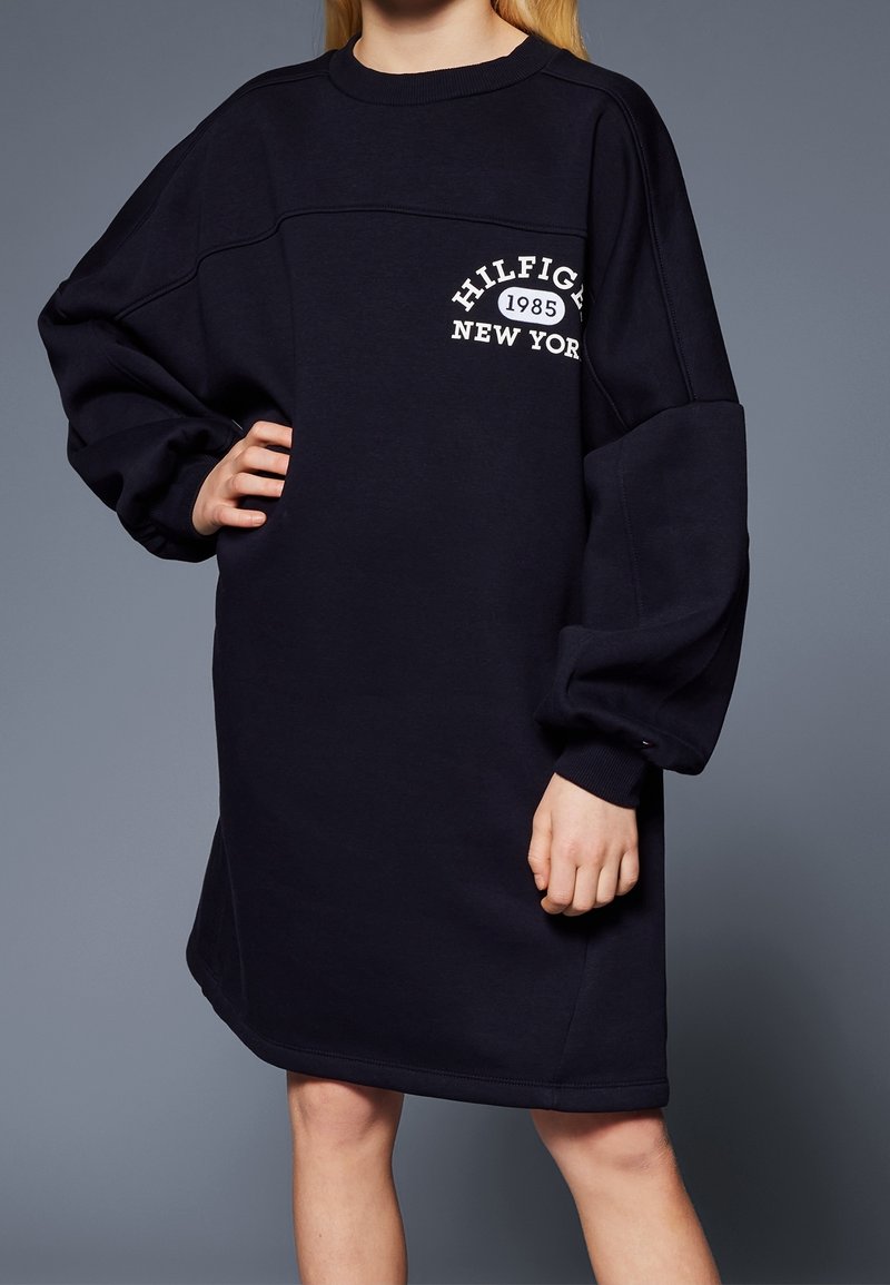 Námořnická modrá oversized mikinová šatová šaty vyrobené z měkkého materiálu mají kulatý výstřih a bílý text loga s vintage designem na hrudi.