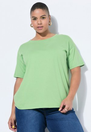 Plus-size woman with short hair wearing gold hoop earrings, yellow eyeliner, a light green t-shirt, and blue jeans, standing against a white wall.
