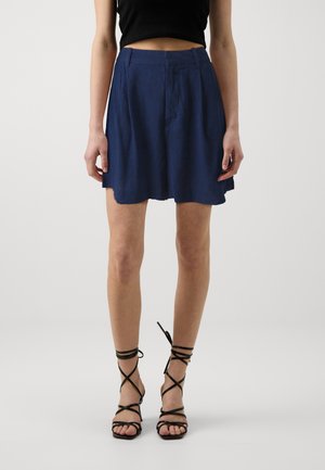 Kvinde iført en høj talje navyblå nederdel med folder, sort crop top og sorte strop-sandaler med høj hæl, stående mod en ensfarvet baggrund.