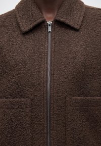 Gros plan d'une veste polaire texturée marron foncé avec fermeture éclair métallique et col portée par une personne à la peau foncée.