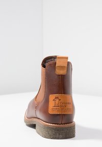Botín de cuero marrón hasta el tobillo con punta redonda, paneles laterales elásticos, suela de goma texturizada y un parche de cuero beige en la parte posterior.