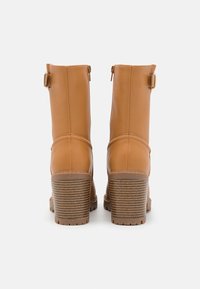 Botas de tobillo marrón claro con un acabado de cuero suave, cremalleras traseras y un tacón apilado texturizado. Presentan acentos de cinturón estrechos en la parte superior.