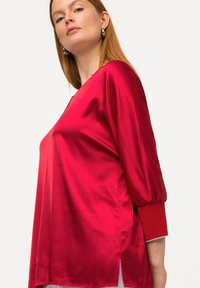 Top en satin rouge avec un décolleté en V, des manches trois-quarts et des fentes sur les côtés. Présente une texture lisse et un léger éclat.