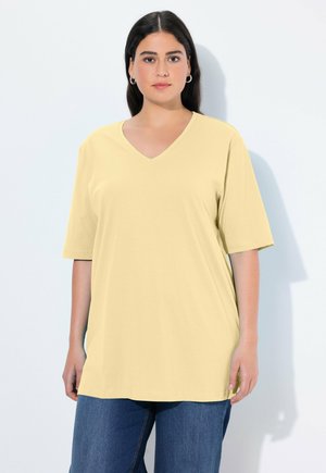 Frau mit dunklen Haaren, die Creolen trägt, ein locker sitzendes, blassgelbes V-Ausschnitt-T-Shirt und blaue Jeans, stehend vor einem einfarbigen hellen Hintergrund.