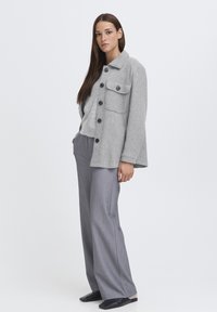 Sivý vlnený kabát s čiernymi gombíkmi a štvorcovou vreckou, navrstvený nad svetlo sivým topom, v kombinácii so sivými širokými nohavicami a čiernymi šľapkami.