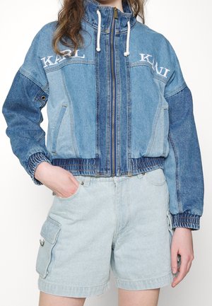 Woman wearing a zip-up patchwork denim jacket with white drawstrings and matching light denim cargo shorts, one hand in pocket.