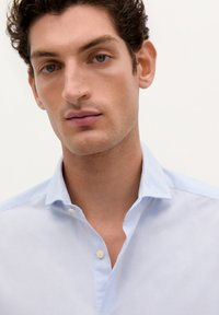 Camisa azul clara de botones con un diseño simple, cuello puntiagudo y una textura sutil. Dos botones en el cuello, acabado limpio.