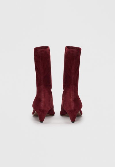 Bottes de cheville en suede bordeaux avec un talon effilé, présentant une texture lisse et un design sans couture. Partie supérieure ouverte avec une légère élasticité au niveau du col.