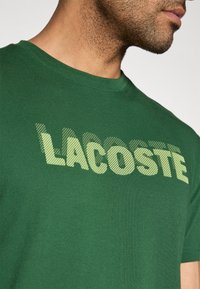 Camiseta de algodón verde con cuello redondo y mangas cortas; presenta un logo "LACOSTE" texturizado en amarillo-verde con una fuente audaz.