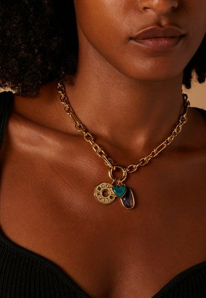 Collier en chaîne dorée avec trois pendentifs : un disque circulaire gravé de "LUCK", un cœur turquoise et une pierre violette, reposant sur une peau texturée.