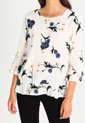 Blouse blanche à imprimé floral bleu et beige, manches et ourlet à volants, portée avec un pantalon noir par une personne debout.