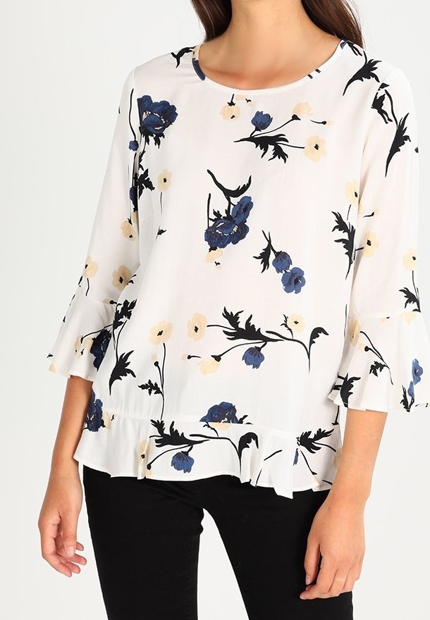 Blouse blanche à imprimé floral bleu et beige, manches et ourlet à volants, portée avec un pantalon noir par une personne debout.
