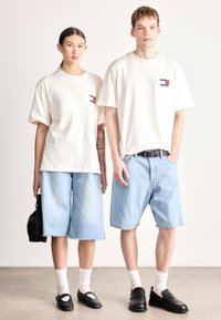 Dois modelos estão lado a lado, vestindo t-shirts bege oversized. A modelo feminina combina a sua com culottes de ganga azul claro e sapatos pretos. O modelo masculino usa calções de ganga e mocassins pretos.