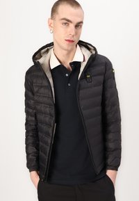 Giacca a vento nera con cappuccio, design trapuntato, chiusura frontale con zip e fodera grigia chiara. Presenta un piccolo logo sul petto e polsini elasticizzati.