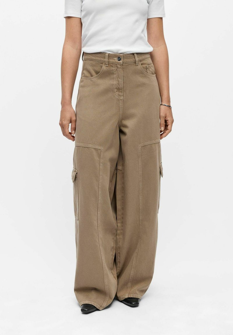 Personne portant un pantalon cargo marron à jambes larges et une chemise blanche à manches courtes, debout devant un fond blanc uni.