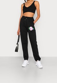 Černé joggingové kalhoty s květinovou výšivkou a stahovací šňůrkou v pase, spárované s černým hladkým crop topem a bílými teniskami.