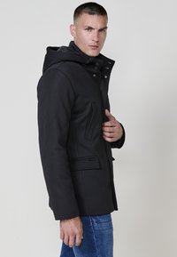 Giacca nera imbottita con cappuccio, dotata di chiusura a zip frontale, bottoni a pressione e tasche laterali; texture liscia e design strutturato.