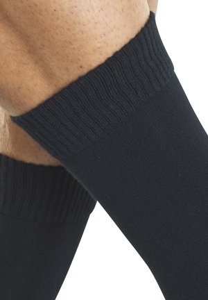 Nahaufnahme von Beinen, die schwarze kniehohe Socken mit gerippten Bündchen vor weißem Hintergrund tragen.