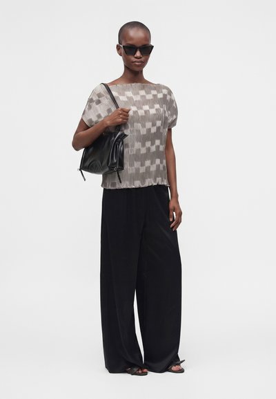 Femme portant un haut à manches courtes transparent à motifs, un pantalon noir large, des sandales noires, des lunettes de soleil et tenant un sac à bandoulière noir.