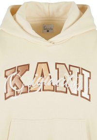 Felpa crema con cappuccio che presenta un grande ricamo "KANI" in tonalità marroni e il testo bianco corsivo “Originator” sul petto. Include una tasca a canguro.