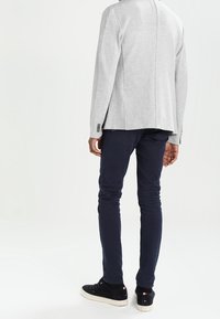 Grauer Strickblazer mit strukturiertem Rücken, kombiniert mit dunkelblauen Slim-Fit-Hosen und schwarzen Sneakern mit weißen Sohlen und Akzenten.