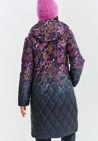 Osoba oblečená v dlouhém prošívaném kabátu s fialovým květinovým gradientem a růžovou pletenou čepicí, pohledem zezadu na neutrálním pozadí.