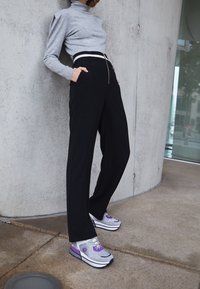 Collo turtleneck grigio, pantaloni neri a vita alta con cerniera frontale, e sneaker grigio/viola con accenti a motivi. Superfici lisce.