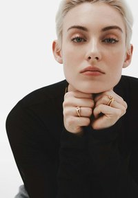 Joven con cabello corto rubio descansa el mentón sobre los puños cerrados, lleva una camiseta negra y anillos dorados con piedras transparentes en los dedos.