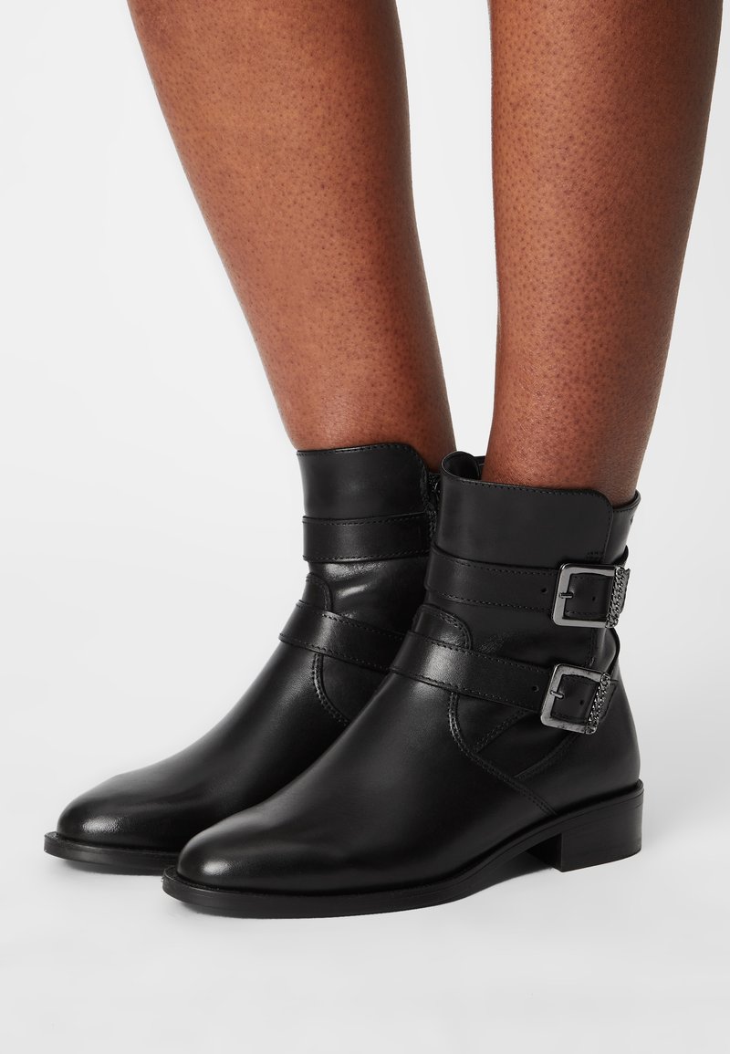 Paire de bottines en cuir noir avec petits talons et double boucle argentée portées sur des jambes brunes sur fond blanc.
